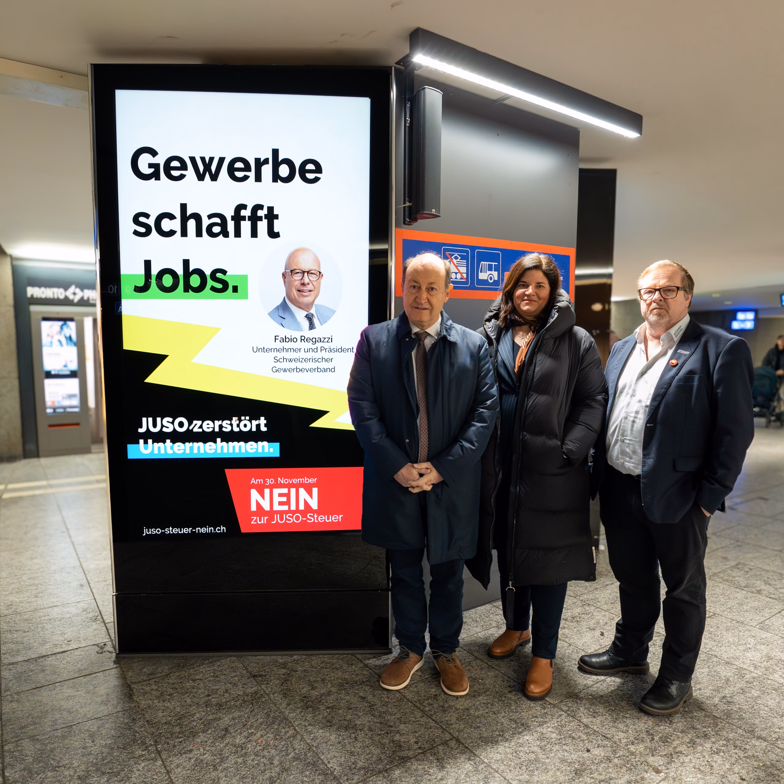 Vertreterinnen und Vertreter der Kampagne gegen die JUSO-Steuer vor einem digitalen Plakat im Bahnhof